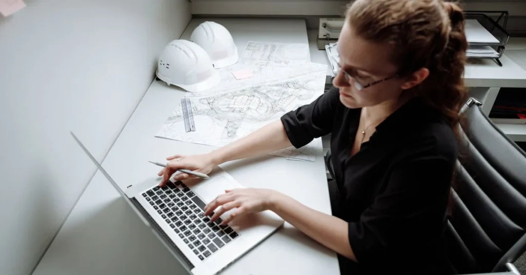 Personne annotant des plans de chantier devant un ordinateur portable, casques posés à côté